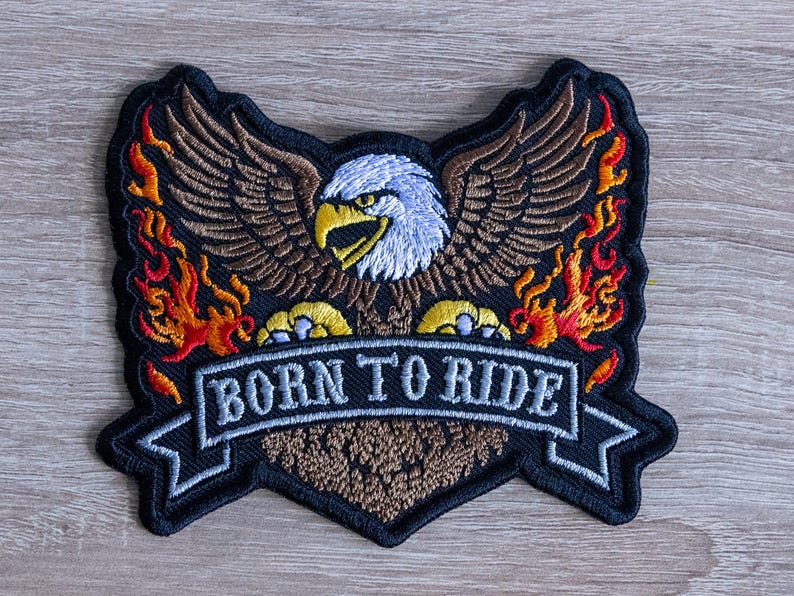 May include: Embroidered patch featuring an eagle with outstretched wings, flames, and the text "BORN TO RIDE" on a banner. The eagle has a white head, brown wings, and yellow talons. The flames are red, orange, and yellow.