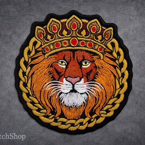 Op de afbeelding: Geborduurde patch met een leeuwenkop met een kroon. De leeuw is oranje en bruin, met een witte snuit en gele ogen. De kroon is goudkleurig met rode accenten en de leeuw is omgeven door een gouden ketting en zwarte rand.
