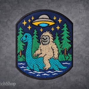Peut inclure: Écusson brodé représentant un monstre du Loch Ness bleu avec un Bigfoot sur son dos, sous un OVNI. L'écusson a un fond bleu foncé avec des arbres, des étoiles et des détails d'eau. L'écusson est de forme unique.