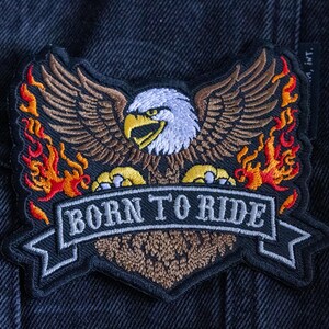 May include: Embroidered patch featuring a detailed bald eagle with outstretched wings, flames, and the text "BORN TO RIDE" on a banner. The patch has a black border and is set against a dark denim background.