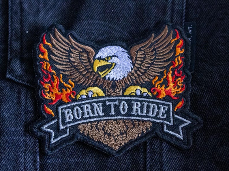 May include: Embroidered patch featuring a detailed bald eagle with outstretched wings, flames, and the text "BORN TO RIDE" on a banner. The patch has a black border and is set against a dark denim background.