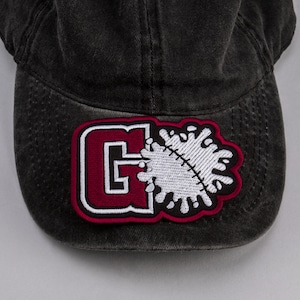 Puede incluir: Una gorra de béisbol negra con un parche bordado en rojo y blanco. El parche presenta una letra "G" y un diseño de salpicadura blanca.