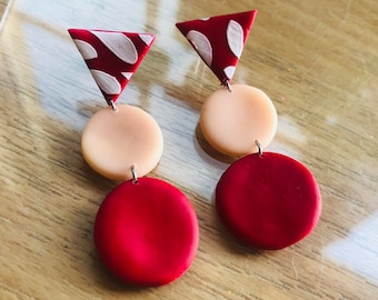 May include: A pair of earrings with three geometric shapes. The top shape is a red triangle with white polka dots. The middle shape is a light peach circle. The bottom shape is a red circle.