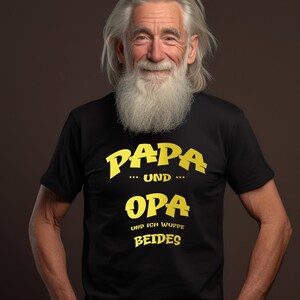 Könnte beinhalten: Ein Mann trägt ein schwarzes T-Shirt mit dem Text "Papa und Opa und ich wuppe beides" in gelben Buchstaben.
