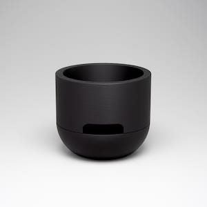 May include: A black cylindrical container with a rounded base and a recessed rectangular opening. The container has a textured surface and a wide opening at the top. The object is set against a neutral background.