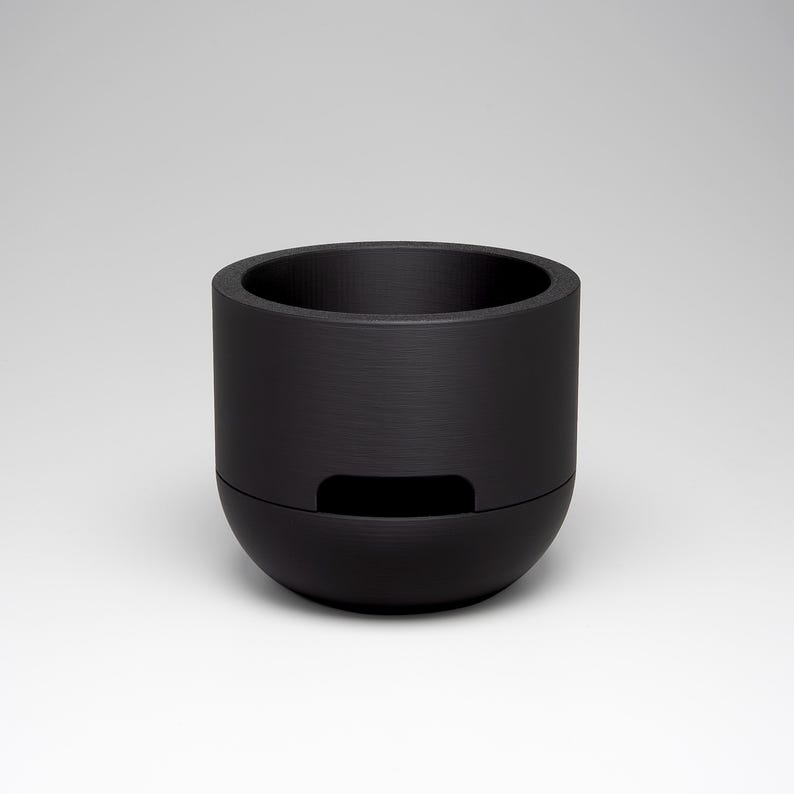 May include: A black cylindrical container with a rounded base and a recessed rectangular opening. The container has a textured surface and a wide opening at the top. The object is set against a neutral background.