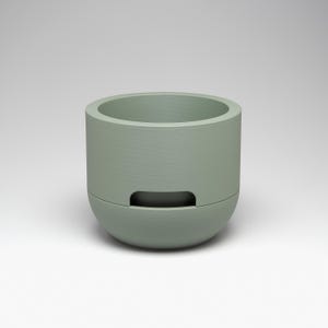 May include: A sage green, cylindrical planter with a rounded base and a rectangular cutout. The planter has a textured surface and a wide opening at the top. The design is modern and minimalist, suitable for small plants or succulents.
