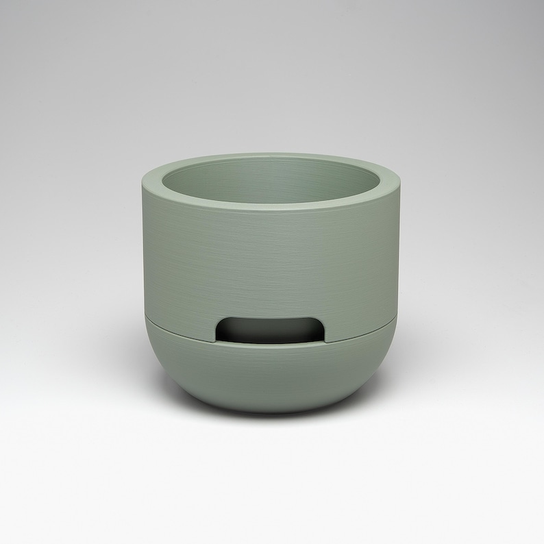 May include: A sage green, cylindrical planter with a rounded base and a rectangular cutout. The planter has a textured surface and a wide opening at the top. The design is modern and minimalist, suitable for small plants or succulents.