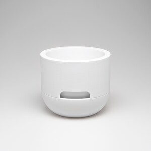 May include: A white, cylindrical container with a rounded base and a recessed handle. The top is open, revealing the interior. The minimalist design features clean lines and a smooth, matte finish. The container is likely made of ceramic or a similar material.