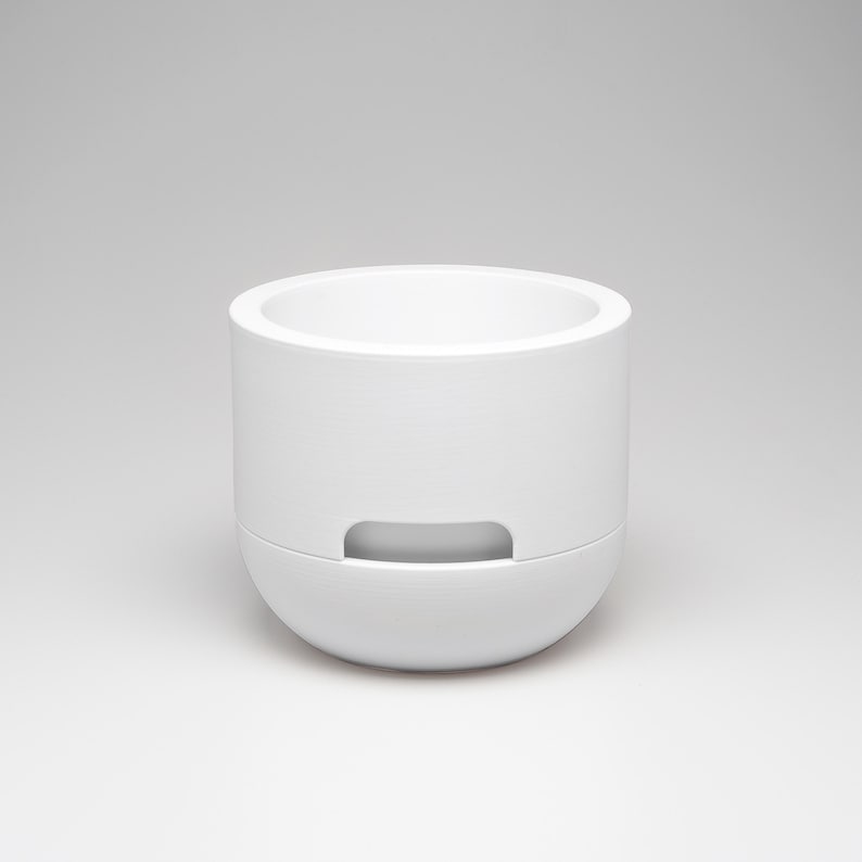May include: A white, cylindrical container with a rounded base and a recessed handle. The top is open, revealing the interior. The minimalist design features clean lines and a smooth, matte finish. The container is likely made of ceramic or a similar material.