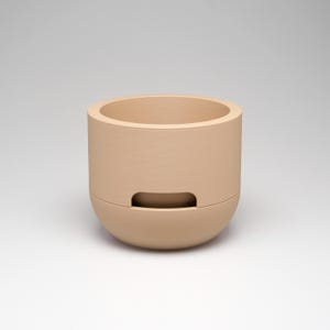 May include: A beige ceramic planter with a cylindrical upper section and a rounded lower section. The planter has a rectangular opening on the front. The neutral color and simple design make it suitable for various decor styles.