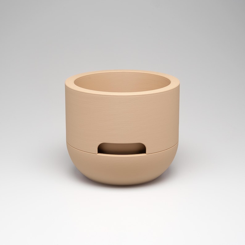 May include: A beige ceramic planter with a cylindrical upper section and a rounded lower section. The planter has a rectangular opening on the front. The neutral color and simple design make it suitable for various decor styles.