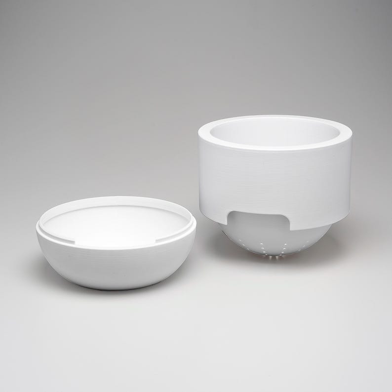 May include: A white ceramic kitchen colander and bowl set. The colander has a cylindrical top with a perforated spherical base. The bowl is a matching white, hemispherical shape. The set is designed for food preparation.