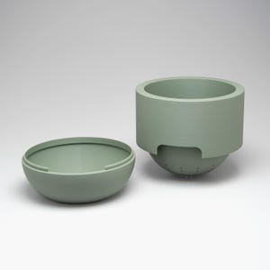 May include: Two-piece sage green kitchenware set. The set includes a round bowl and a cylindrical container with a perforated base. The container has a wide rim and a curved handle-like design. The bowl is open and has a rounded shape.