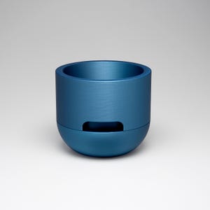 May include: A cylindrical, blue container with a rounded base and a recessed rectangular opening. The container has a brushed metal texture and a wide opening at the top. The object is centered against a white background.
