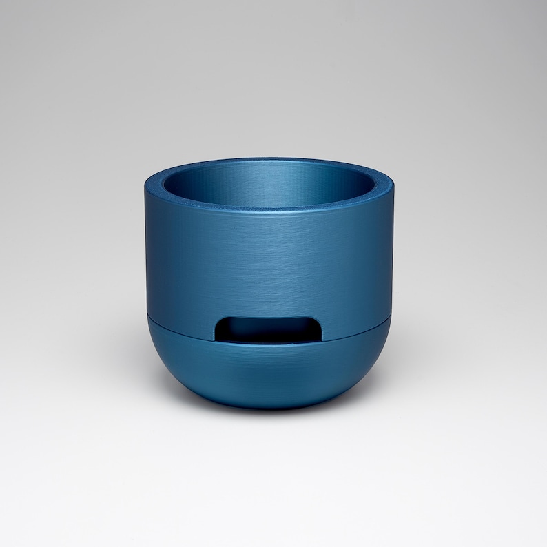 May include: A cylindrical, blue container with a rounded base and a recessed rectangular opening. The container has a brushed metal texture and a wide opening at the top. The object is centered against a white background.