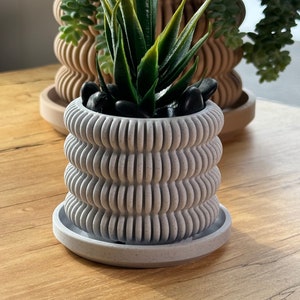 Puede incluir: Una maceta de cerámica blanca con un diseño texturizado ondulado. La maceta tiene un platillo a juego y está llena de piedras negras y una pequeña planta suculenta verde.