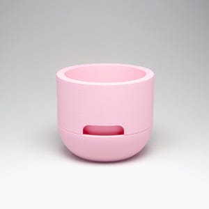 May include: A light pink, cylindrical planter with a rounded base. The planter has a recessed handle on the lower section and a wide opening at the top. The interior is also light pink. The planter is designed for indoor or outdoor use.