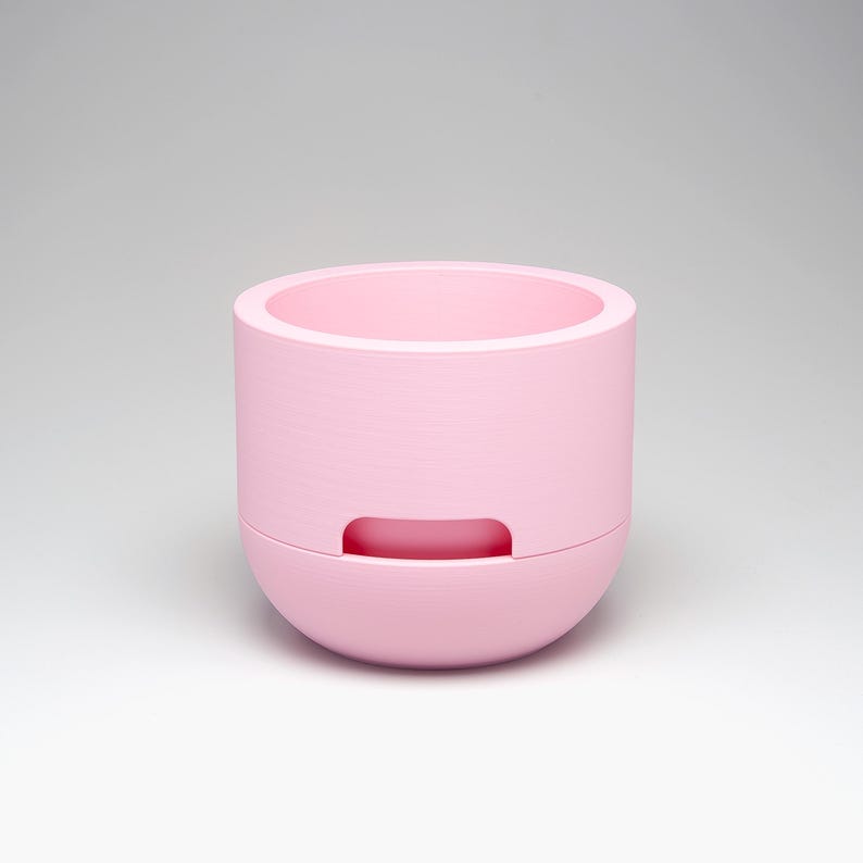 May include: A light pink, cylindrical planter with a rounded base. The planter has a recessed handle on the lower section and a wide opening at the top. The interior is also light pink. The planter is designed for indoor or outdoor use.