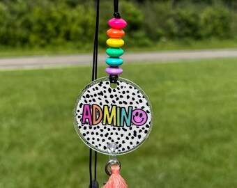Puede incluir: Una insignia redonda de ADMIN con un fondo de lunares blancos y negros, una borla rosa y un acento de cuentas de arcoíris. La palabra "ADMIN" está en letras rosas con un emoji de carita sonriente. La insignia cuelga de un cordón negro.