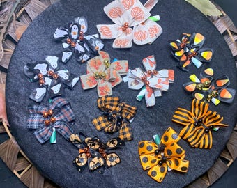 May include: A collection of decorative hair bows in various Halloween-themed designs. The bows feature patterns like pumpkins, ghosts, and candy corn, with accents of orange, black, and white. Each bow is embellished with small beads.