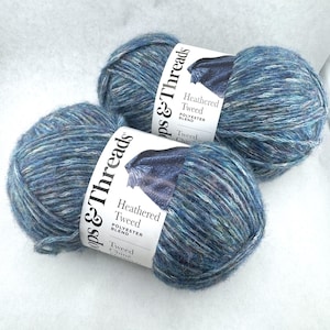 May include: Two skeins of Loops & Threads Heathered Tweed polyester blend yarn in a variegated blue colorway. Each skein features a white paper label with black text detailing the brand name, product line, and material composition.