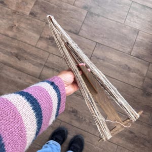 May include: A wooden yarn swift with a beige and white yarn wound around it. The swift is being held by a person wearing a pink and blue striped sweater.