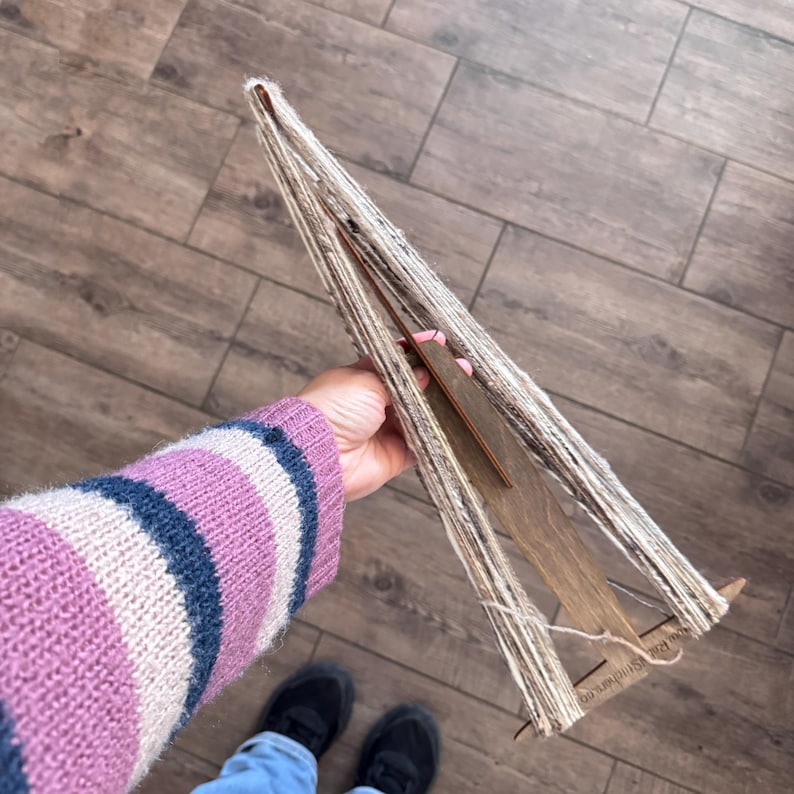 May include: A wooden yarn swift with a beige and white yarn wound around it. The swift is being held by a person wearing a pink and blue striped sweater.