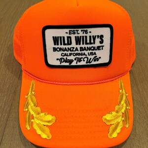 May include: Orange trucker hat with a black and white patch that reads "EST. 76 - WILD WILLY'S BONANZA BANQUET CALIFORNIA, USA "Play To Win" ". The hat has two gold embroidered feathers on the brim.