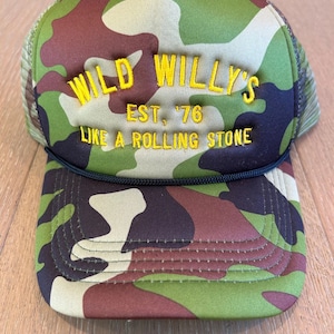 Puede incluir: Una gorra de camionero de camuflaje con el texto "WILD WILLY'S EST. '76 LIKE A ROLLING STONE" bordado en amarillo.