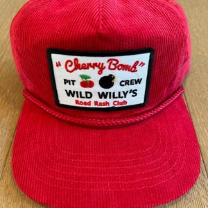 May include: Red corduroy baseball cap with a black and white patch that reads "Cherry Bomb" "Pit Crew Wild Willy's Road Rash Club".