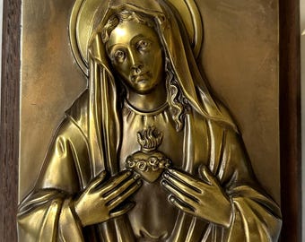 May include: A gold-toned metal relief sculpture of the Virgin Mary with her hands clasped over her heart. The heart is adorned with a crown of thorns and a flame.