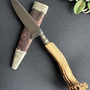 Pode incluir: Uma faca de caça vintage com uma bainha de couro marrom e um cabo de chifre de veado. A lâmina é de aço e tem uma ponta pontiaguda.