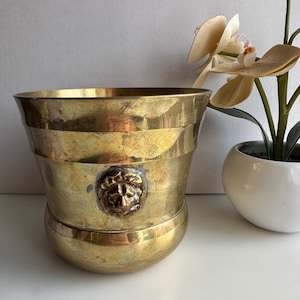 May include: A vintage brass planter with a decorative lion's head emblem. The planter has a wide, fluted body and a rounded base. The brass has a warm, aged patina. The planter is empty.