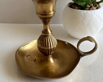 Puede incluir: Un candelero de latón con una base redonda y un asa. El candelero tiene un diseño decorativo en la base y una copa para la vela.