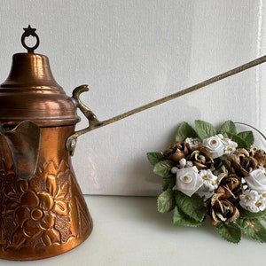 May include: A copper Turkish coffee pot with an ornate floral design and a long handle. The pot is sitting on a white surface with a bouquet of white and gold flowers.