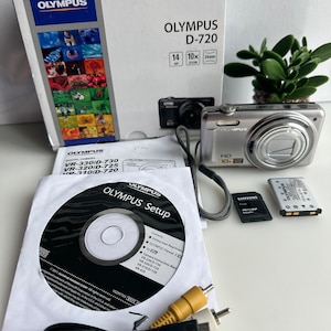 Peut inclure: Un appareil photo numérique Olympus D-720 argenté avec un zoom 10x et un capteur 14MP. L'appareil photo est emballé dans une boîte blanche avec le logo Olympus et les spécifications de l'appareil photo. L'appareil photo est également présenté avec un CD, un câble USB, un câble RCA jaune, une carte mémoire et une batterie.