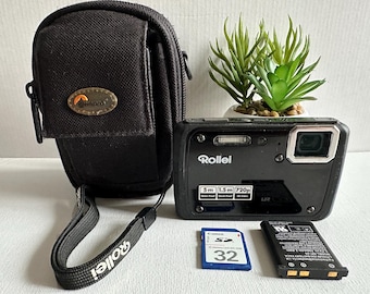 Puede incluir: Una cámara digital negra con una lente plateada y una funda de transporte negra. La cámara tiene la palabra "Rollei" impresa en la parte delantera. La cámara está sobre una superficie blanca con una pequeña planta en maceta en el fondo.