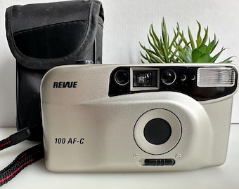 Puede incluir: Cámara plateada Revue 100 AF-C con lente y flash negros, acompañada de una funda negra. La cámara tiene una correa negra con detalles rojos. Una pequeña planta verde está encima de la cámara.