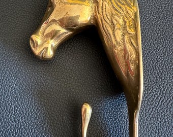 May include: A brass hook in the shape of a horse's head. The hook is a golden color and features detailed craftsmanship, including the horse's mane and facial features. The hook curves downward, ending in a rounded tip.