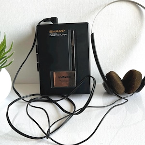 May include: Black Sharp stereo cassette player with X-Bass and headphones. The player has the Sharp logo and text. The headphones have brown ear pads. A small potted plant is in the background.