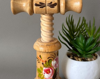 Puede incluir: Un sacacorchos de madera con un diseño floral pintado a mano. El sacacorchos tiene un eje en espiral y dos mangos de madera. El diseño floral presenta rosas rosas, blancas y verdes.