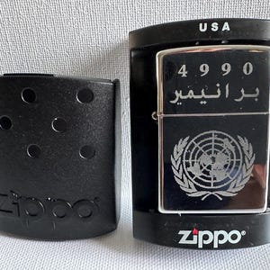 May include: A silver Zippo lighter with the United Nations emblem and Arabic text, displayed in a black case. The lighter has the number 4990 engraved on it. A separate black case with the Zippo logo is also present.