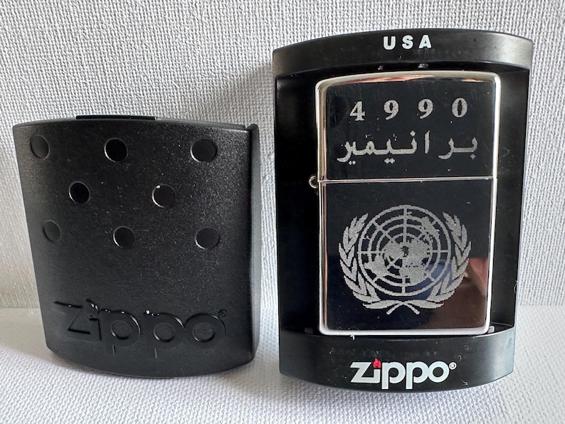 May include: A silver Zippo lighter with the United Nations emblem and Arabic text, displayed in a black case. The lighter has the number 4990 engraved on it. A separate black case with the Zippo logo is also present.