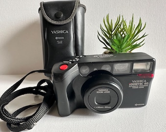 Puede incluir: Cámara Yashica Zoomtec 60 negra con objetivo zoom, correa y funda a juego. La cámara tiene un botón rojo y el texto "Yashica Zoomtec 60 Automatic Focusing Power Zoom". La funda tiene el logotipo de Yashica.