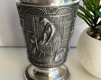Puede incluir: Un tanque de peltre plateado con un diseño en relieve de una mujer amasando masa. El tanque tiene una base redonda y una parte superior acampanada. La inscripción "Beim Teigmengen" es visible en la base.