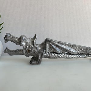 May include: A silver-colored dragon-shaped nutcracker. The dragon has detailed wings and an open mouth, revealing teeth. The nutcracker is lying on a white surface, with a small potted plant in the background.
