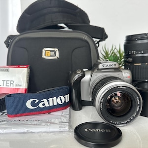 May include: A silver Canon EOS 300 SLR camera with a 28-90mm lens, a black camera bag, a Tamron UV filter, a spare Tamron lens, a blue and red Canon neck strap, a lens cap, and a camera body cap, along with a printed instruction manual.