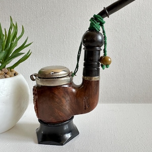 May include: A vintage tobacco pipe with a dark brown, curved bowl and a silver-colored lid. The pipe has a black stem and is adorned with a green cord and a wooden bead. It rests on a black stand, showcasing a classic design.
