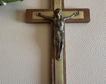Puede incluir: Un crucifijo de madera con una figura de bronce de Jesucristo. La cruz es de madera marrón oscuro con una placa de metal color latón. Las letras "INRI" son visibles en la parte superior. La figura tiene los brazos extendidos.